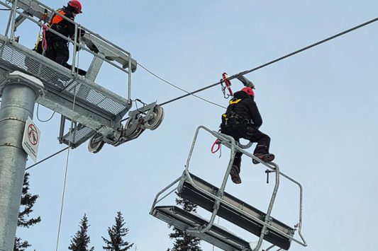 Two workers wearing helmets, harnesses, and winter gear are performing maintenance on a ski lift. One person stands on a small metal platform attached to a lift tower, while the other is suspended from the lift cable above an empty chair. Snow covered evergreen trees fill the background, and the scene takes place outdoors in winter conditions.