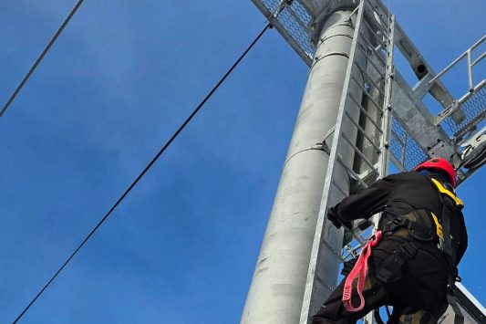 A worker wearing a helmet, harness, and protective clothing is climbing a tall metal tower structure. The tower has a fixed vertical ladder and cables running alongside it. The worker is secured with fall protection gear, and the sky behind them is clear and bright.